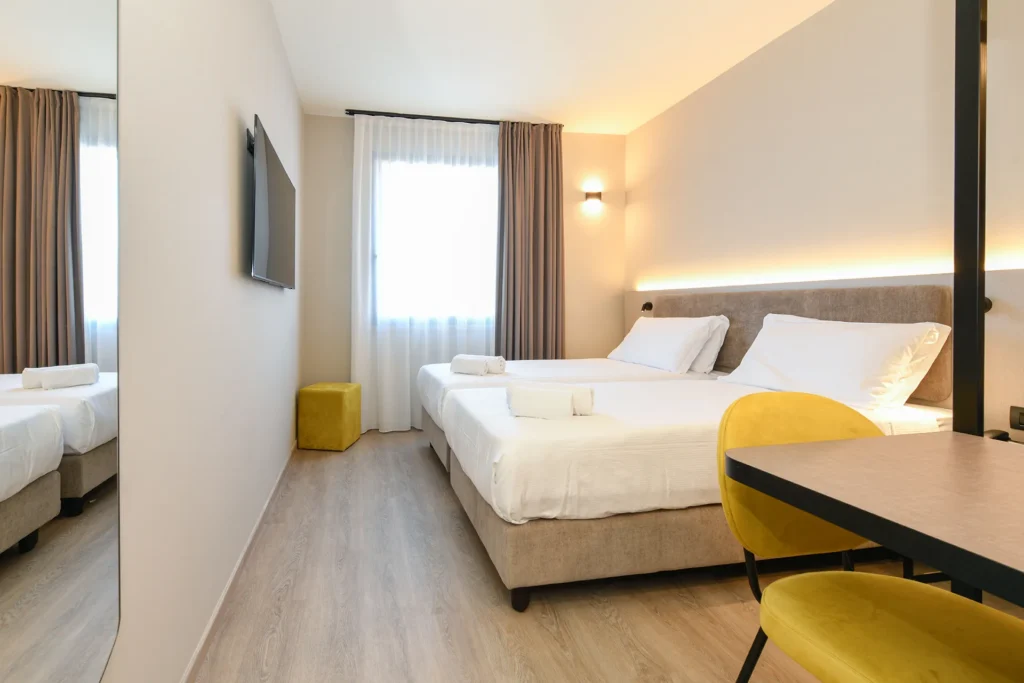 Camera matrimoniale luminosa con letto queen size, finestra con tende chiare e sedia gialla accanto alla scrivania.